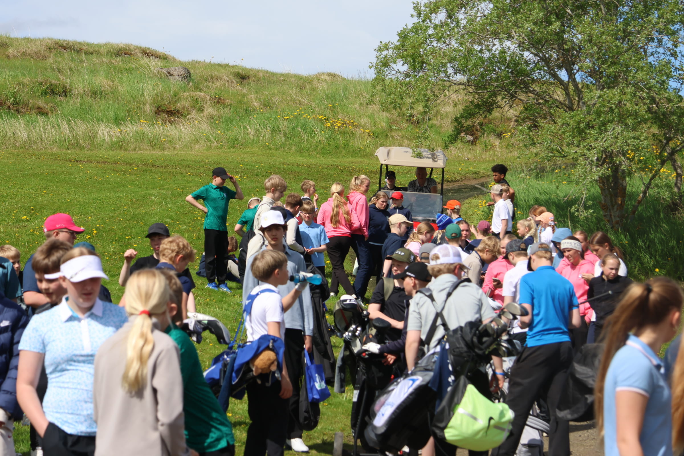 Group of children with backpacks gathered on a sunny grassy field, some talking near a small cart and a hill in the background.
