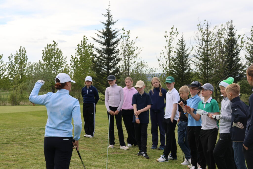 499A0425 - Golfsamband Íslands Instructor in a light blue jacket demonstrates with a golf club to a line of children on a grassy field outdoors, trees in the background.