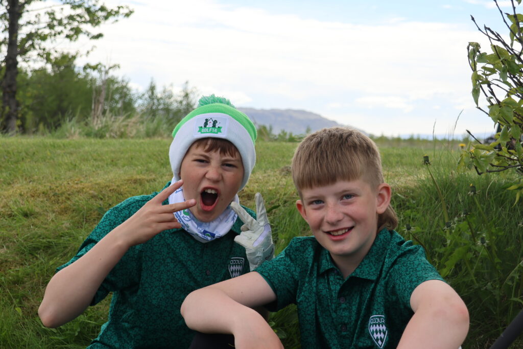 499A0448 - Golfsamband Íslands Two children pose for a selfie in a grassy field, the boy on the left making a peace sign with his mouth open in a playful expression.