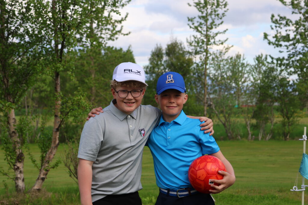 499A0475 - Golfsamband Íslands Two boys outdoors with trees in the background, arms around each other, one holding a red soccer ball central to the frame.