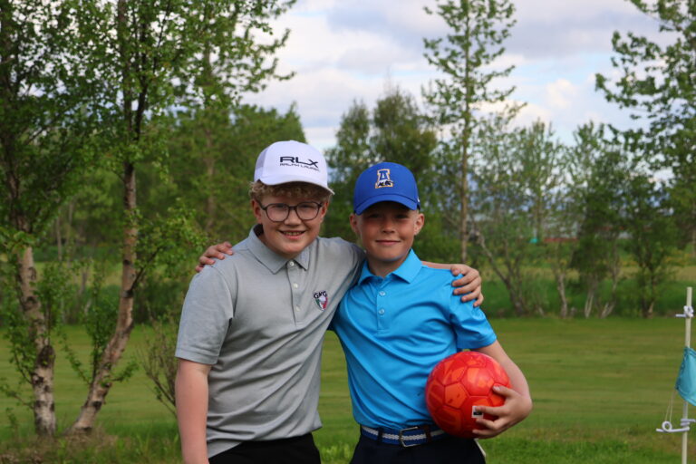 Two boys outdoors with trees in the background, arms around each other, one holding a red soccer ball central to the frame.