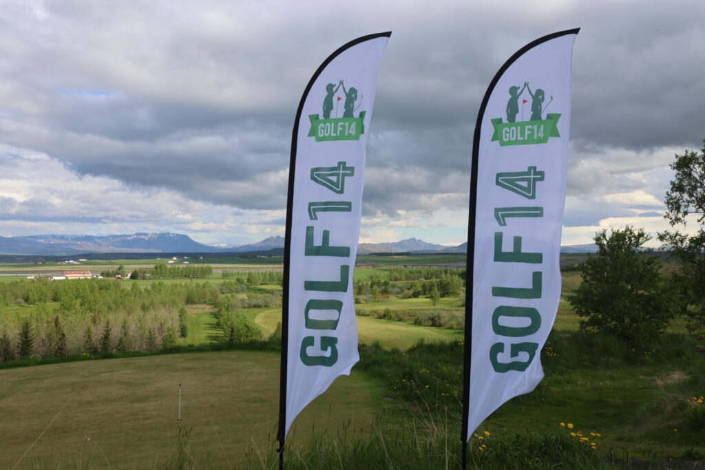 499A0487 - Golfsamband Íslands Two tall white banners with a green 'GOLF14' logo and golfer silhouettes stand in a grassy field against a cloudy, mountainous backdrop.
