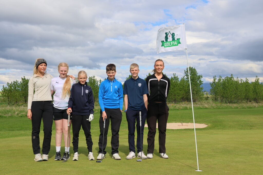 499A0504 - Golfsamband Íslands Six young golfers pose on a green golf course with a Hole flag reading 'GOLF14' under a cloudy sky.