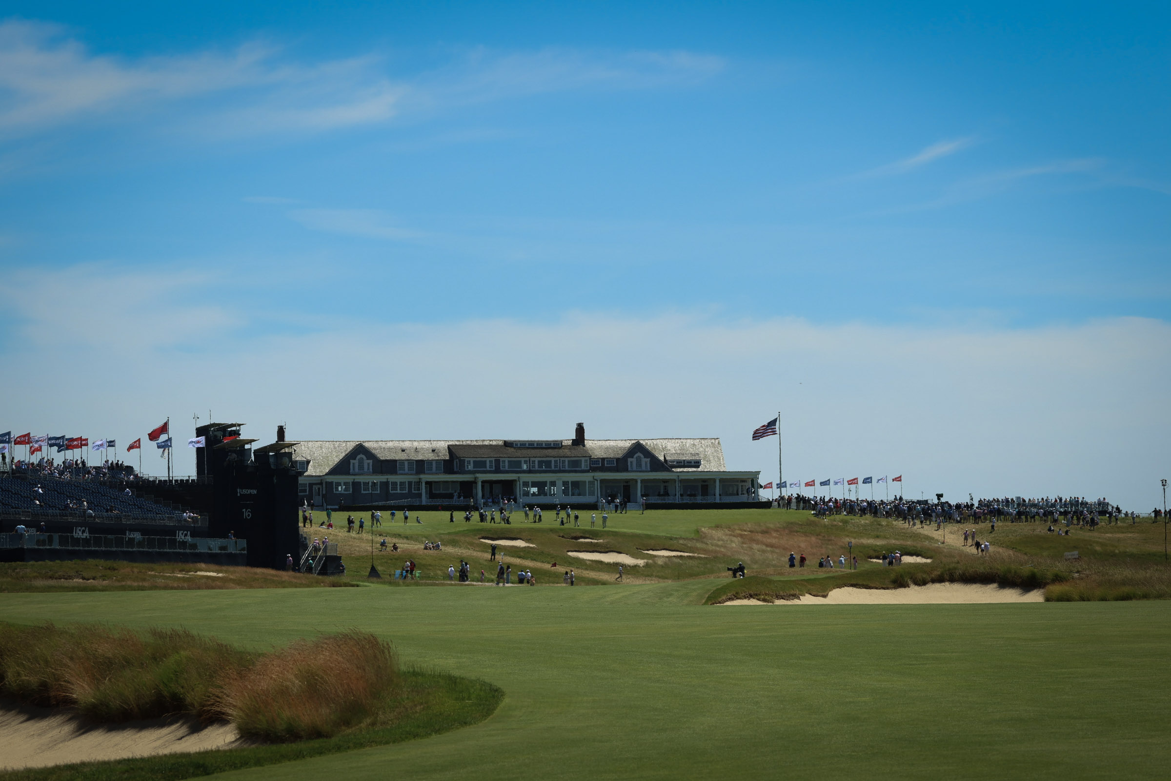Golf course with a long grandstand lined by flags, a clubhouse in the distance, and a clear blue sky above; spectators along the course.