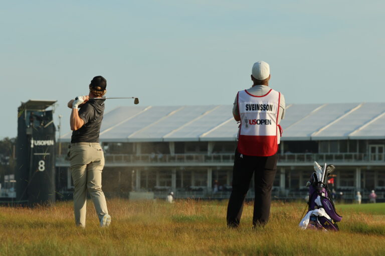 Two golfers on a sunny course: one in a gray shirt swinging a club, the other in a white cap and red-and-white vest labeled SVEINSSON, with golf bags nearby.