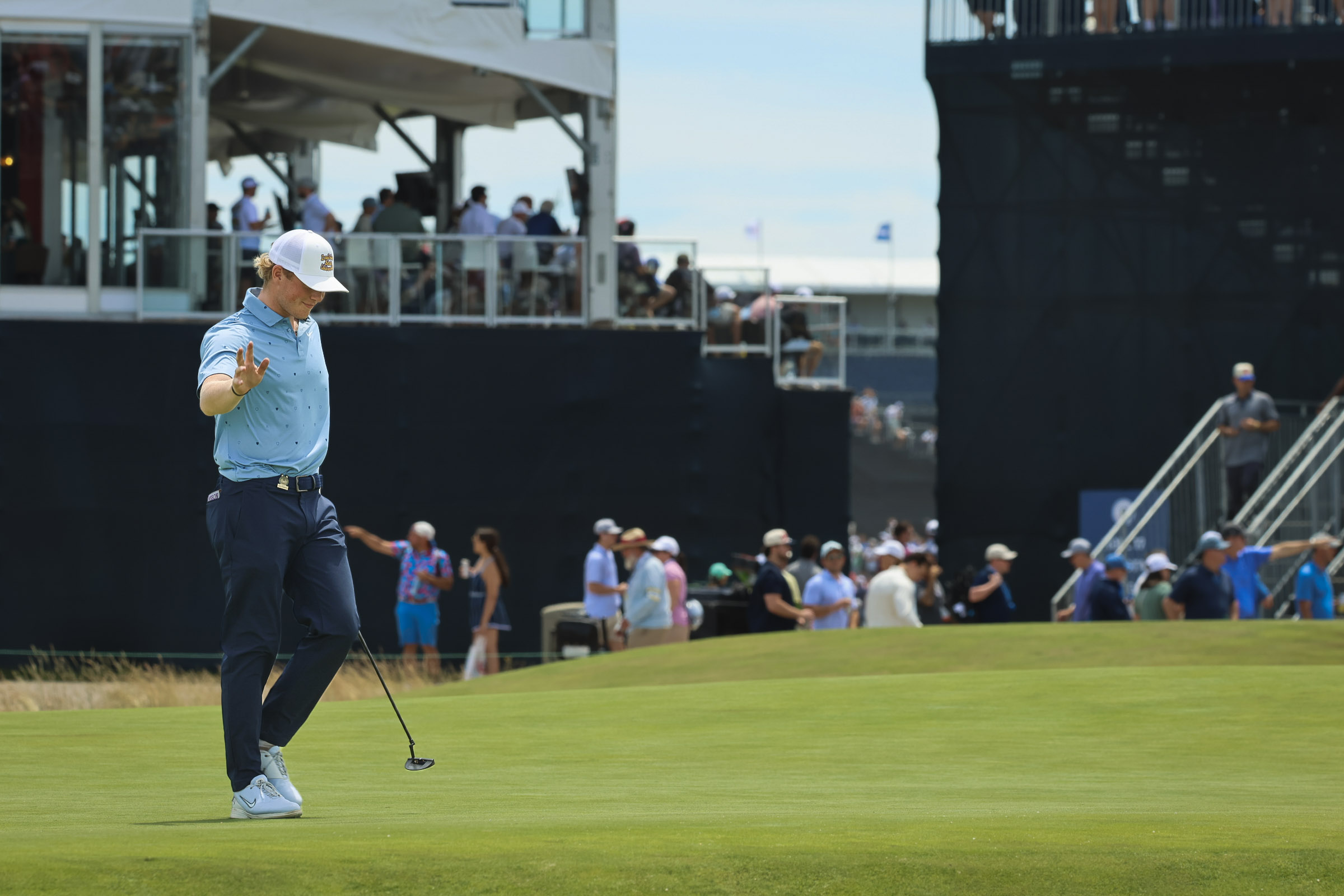 Golfer in a light blue polo and white cap on a green field with a crowd and grandstands in the background.