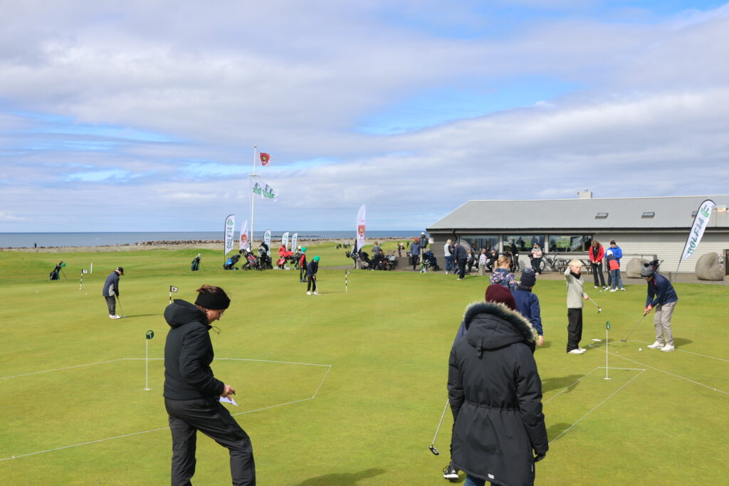 499A9099 - Golfsamband Íslands Group of golfers practicing on a coastal putting green near a modern clubhouse, with flags and spectators in the background.