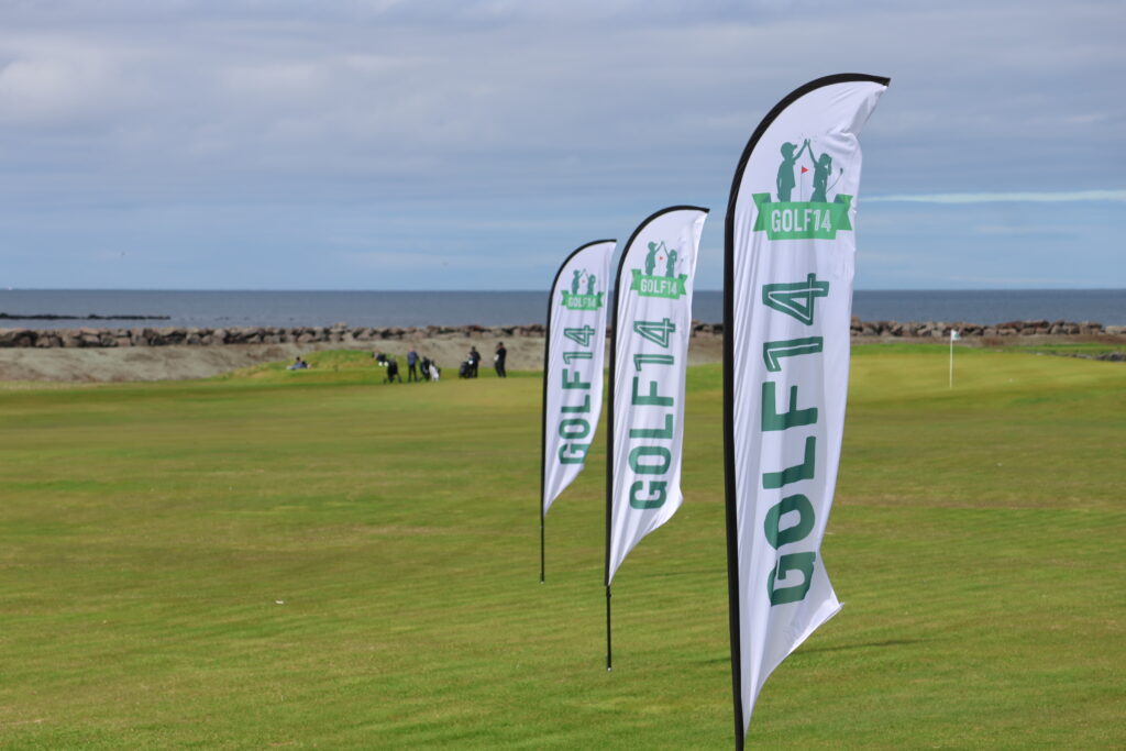 499A9103 - Golfsamband Íslands Row of white flags with green 'GOLF14' logos fluttering on a grassy coastal course, distant people visible near a rocky breakwater with the sea in the background.