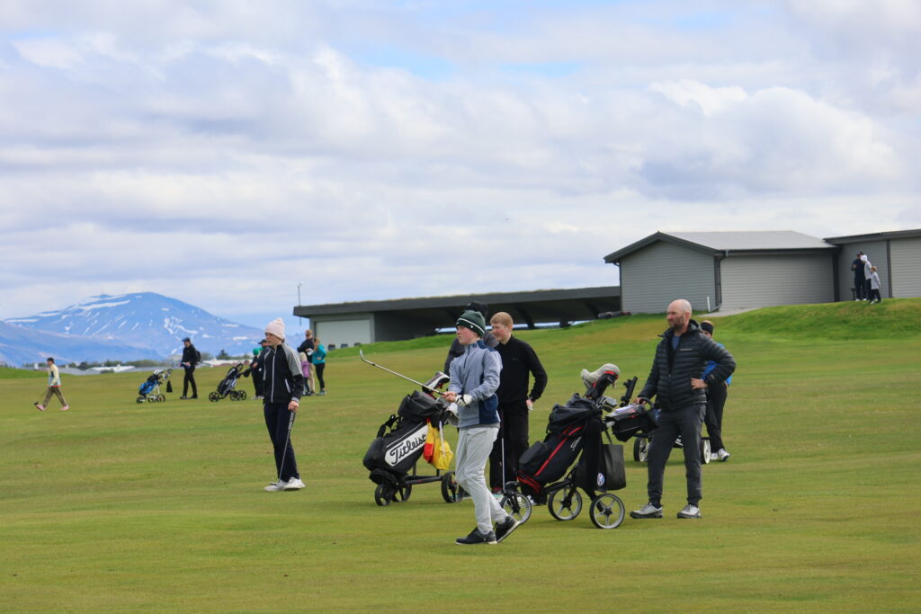 499A9114 - Golfsamband Íslands Group of golfers pushing carts and carrying bags on a grassy golf course, with mountains in the distance and a building to the right.