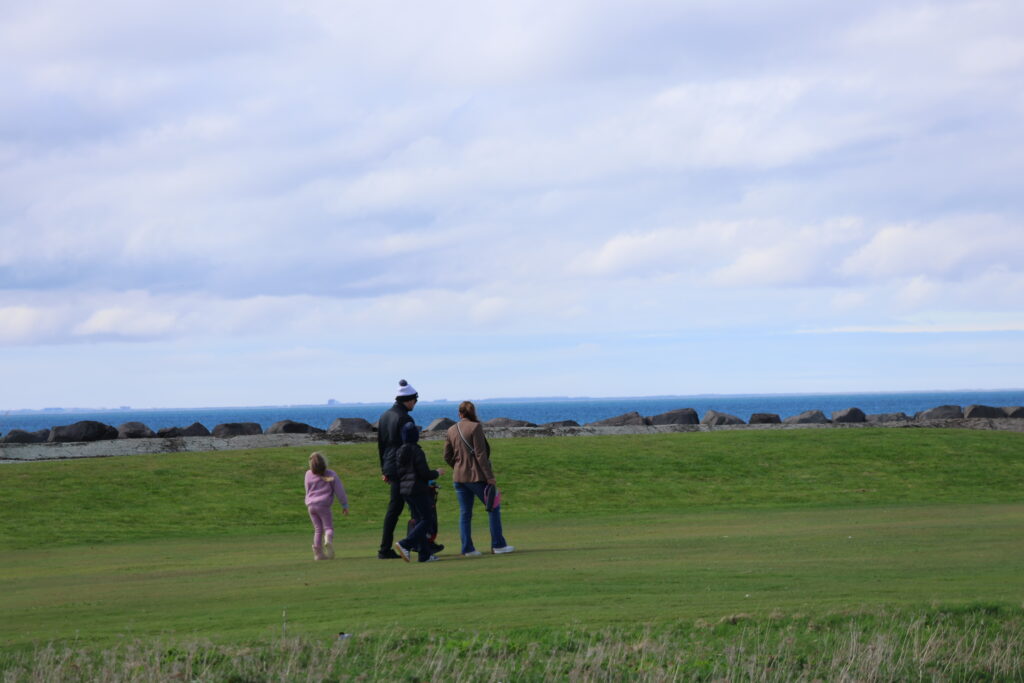 499A9132 - Golfsamband Íslands Group of people walking on a grassy coastal park with a rocky breakwater and the sea in the distance under a cloudy sky.