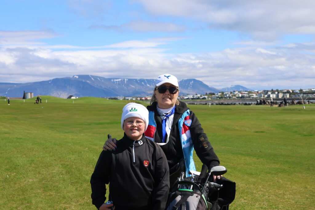 499A9137 - Golfsamband Íslands Two people posing on a grassy field with golf clubs, mountains in the background and a town in the distance.