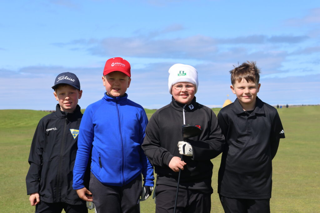 499A9139 - Golfsamband Íslands Four boys in golf attire stand together on a grassy golf course under a blue sky, one holding a golf club.