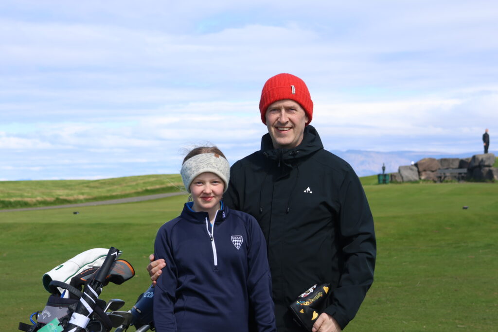 499A9142 - Golfsamband Íslands Two people posing outdoors on a grassy field with a golf bag in the foreground; man in a red knit hat and black jacket, woman in a navy jacket closest to camera.