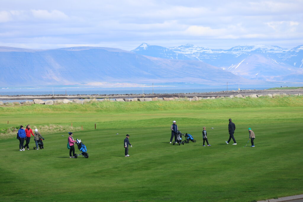 499A9145 - Golfsamband Íslands Golfers with bags and push carts on a bright green coastal golf course, mountains visible in the distance.