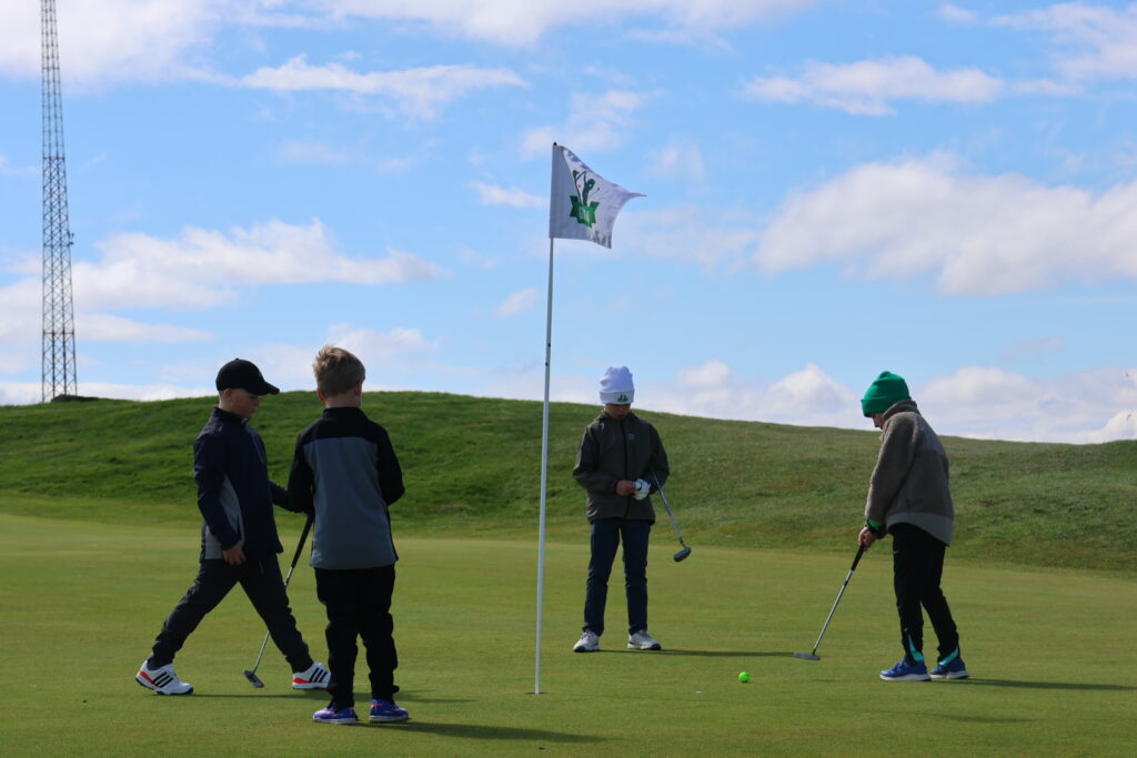 499A9150 - Golfsamband Íslands Group of young golfers on a sunny golf course green near the hole with a flag and golf clubs nearby