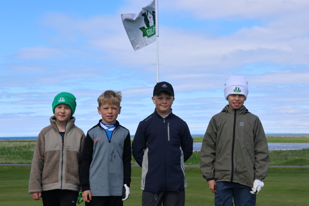 499A9155 - Golfsamband Íslands Five young golfers pose together outdoors on a golf course, with a flagpole and blue sky behind them.