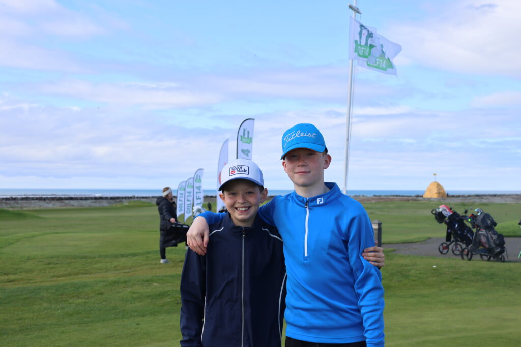 499A9165 - Golfsamband Íslands Two boys posing with arms around each other on a golf course, blue jackets and caps, flags fluttering in the background by the sea.