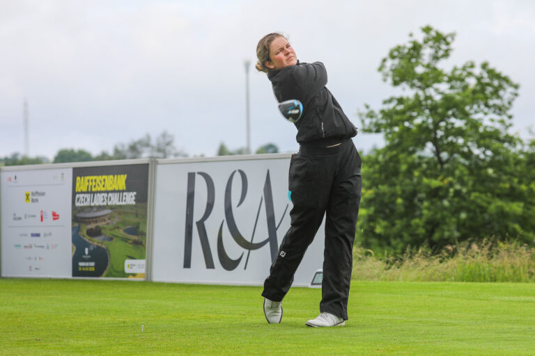 Woman in black athletic wear mid-swing with a club on a grassy field, event banners in the background.