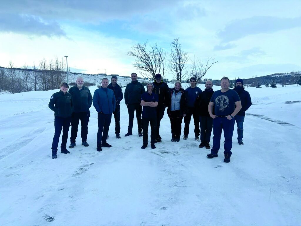 Group of people standing together on a snow-covered field, with leafless trees and hills in the background.