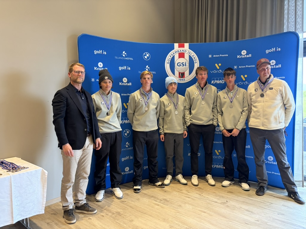 Seven people pose for a medal ceremony in front of a blue sponsor backdrop; six youths in gray jackets with medals and one man in a blazer on the left.