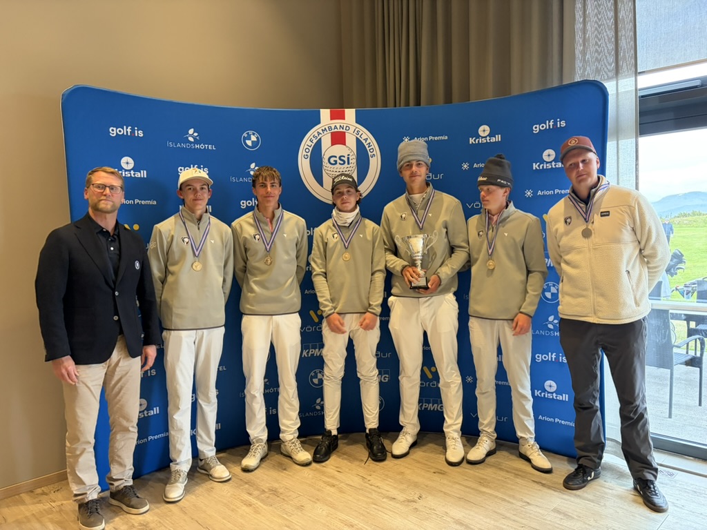 Group of eight people posing in front of a blue sponsor backdrop; all wear medals, gray sweatshirts, and casual pants, with one person holding a trophy in the center.