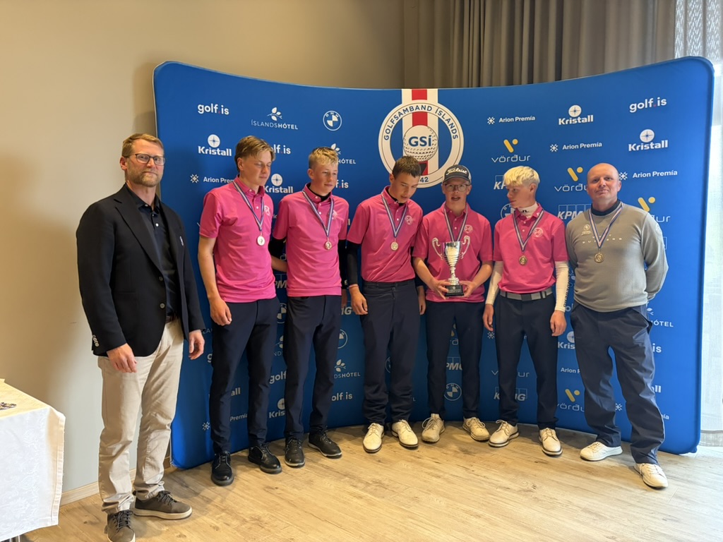Young golfers in pink shirts with medals pose on stage beside a trophy and two coaches in front of a blue sponsor backdrop.