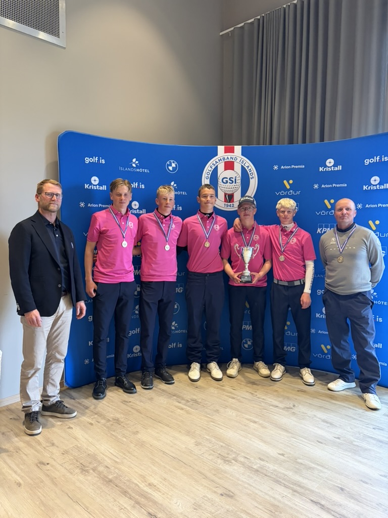 Group of seven men posing in a row; five wearing pink shirts with medals, two in casual outfits, one holding a trophy in front of a blue sponsor backdrop.