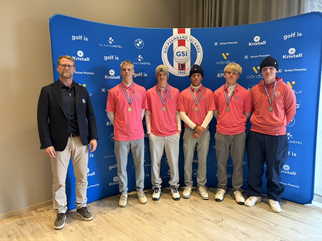 Six young athletes wearing pink shirts and medals stand with a man in a blazer against a blue sponsor backdrop full of logos.