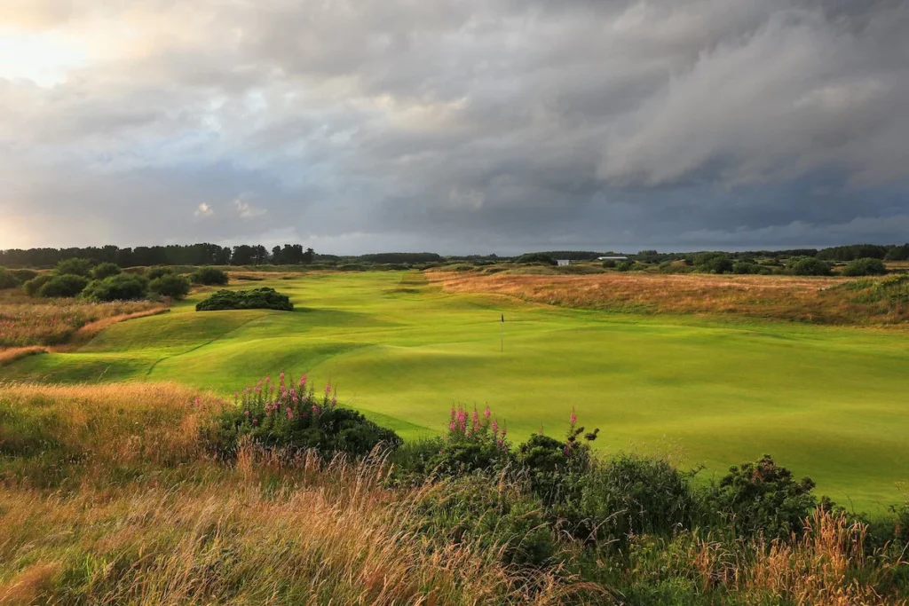 Open golf course fairway under dramatic stormy sky with dark clouds and golden field edges in the distance.