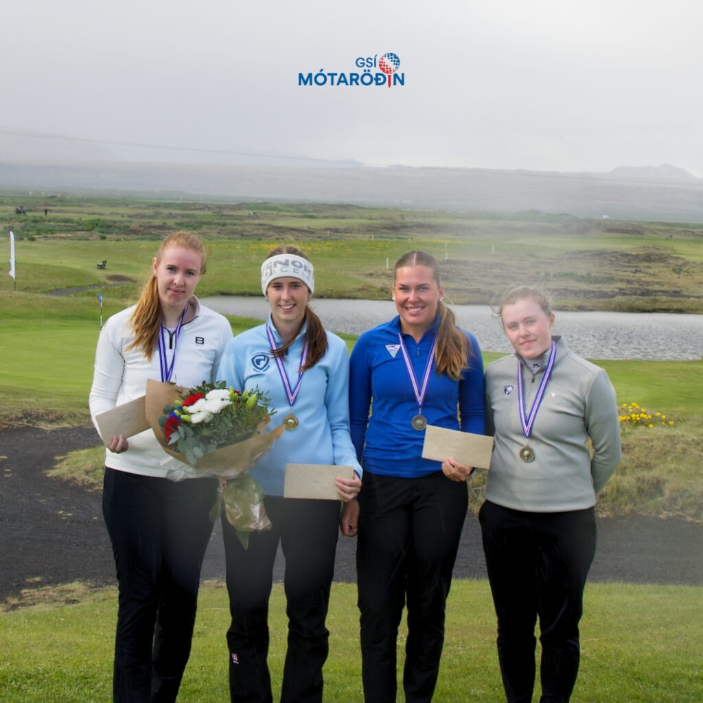 Heading - 2026-06-07T180223248 - Golfsamband Íslands Four female athletes stand on a golf course, wearing medals and holding envelopes; one holds a bouquet, with the MÓTARÖDIN logo above in the sky/air.”