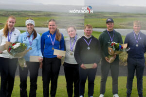 Seven young athletes stand in a row wearing medals, some holding bouquets and envelopes, outdoors with a grassy field and water behind them, logo above.