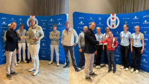 Two groups of young athletes shake hands on a podium as medals and trophies are awarded in front of a blue sponsor wall.