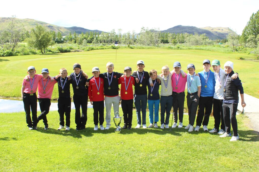 IMG_2833 1 - Golfsamband Íslands Group of children wearing medals posing on a golf course with a trophy in front of them on the grass.