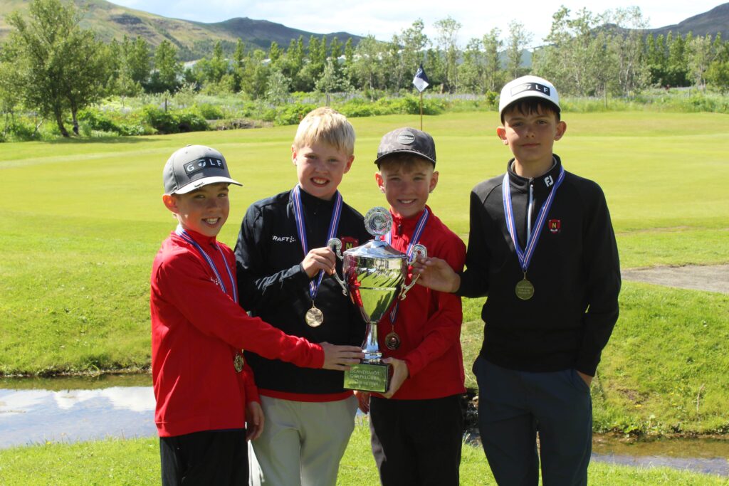 IMG_2837 1 - Golfsamband Íslands Four young golfers pose with a large trophy on a sunny golf course, medals around their necks.