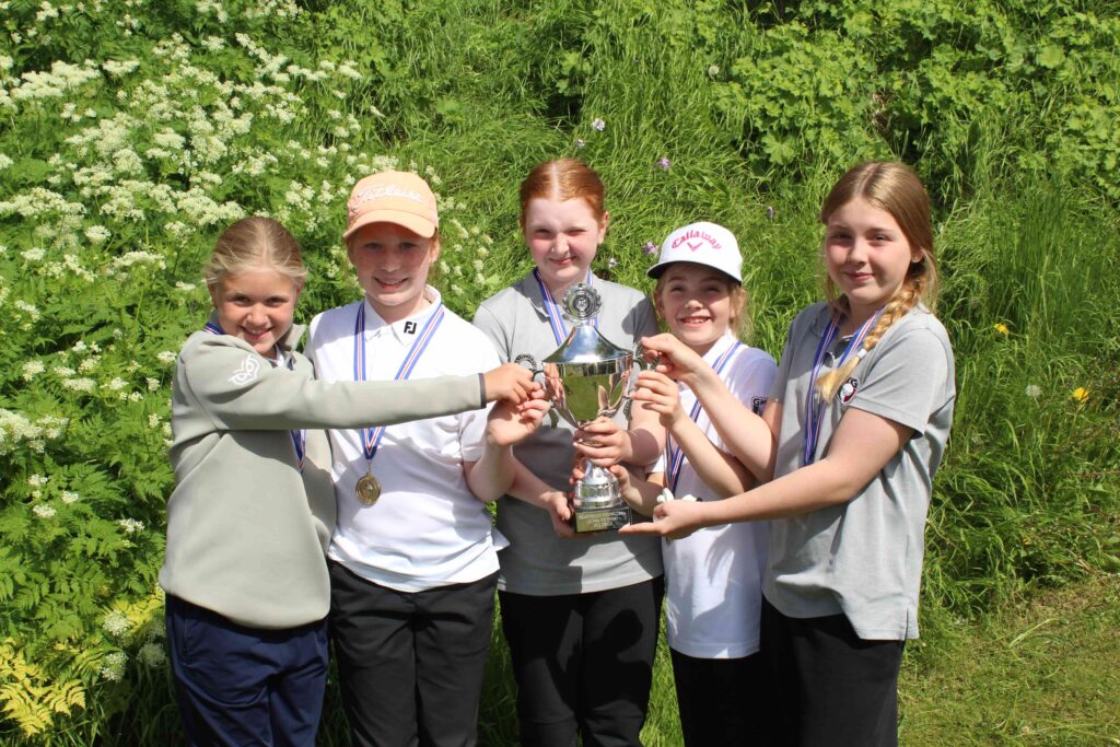 IMG_2841 1 - Golfsamband Íslands Group of smiling girls in casual sportswear posing with a large trophy outdoors, medals around their necks, green foliage in the background.
