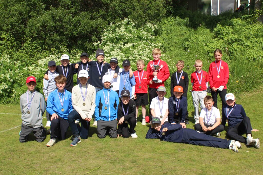 IMG_2847 1 - Golfsamband Íslands Group of children posing outdoors with medals around their necks; one boy in red holds a trophy on a grassy field.