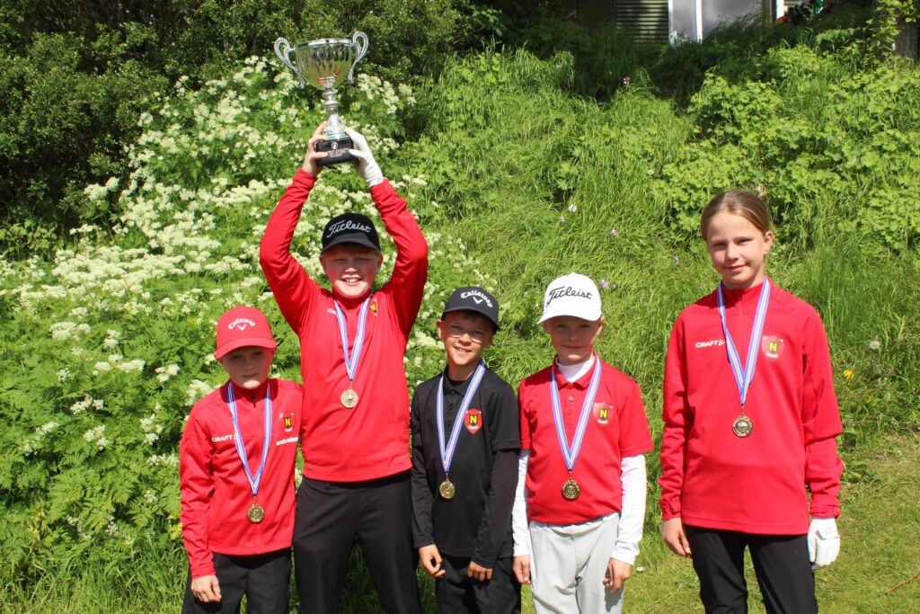IMG_2850 1 - Golfsamband Íslands Young golfers in red uniforms pose outdoors with a silver trophy and medals, celebrating a win.