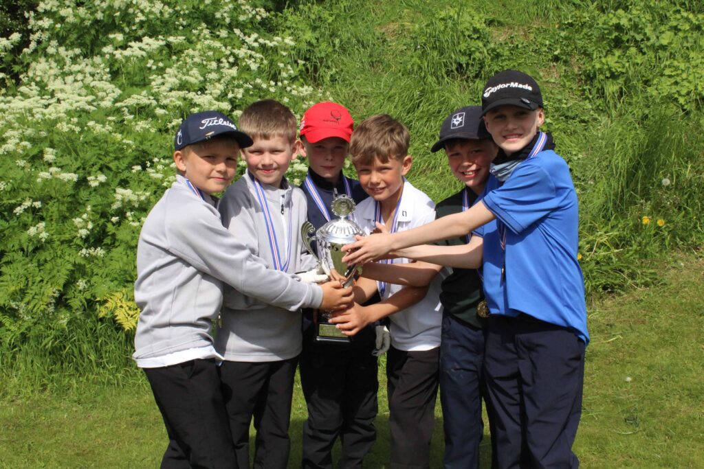 IMG_2864 1 - Golfsamband Íslands Six boys outdoors wearing caps and medals, holding a large silver trophy together in front of green plants and white flowers.