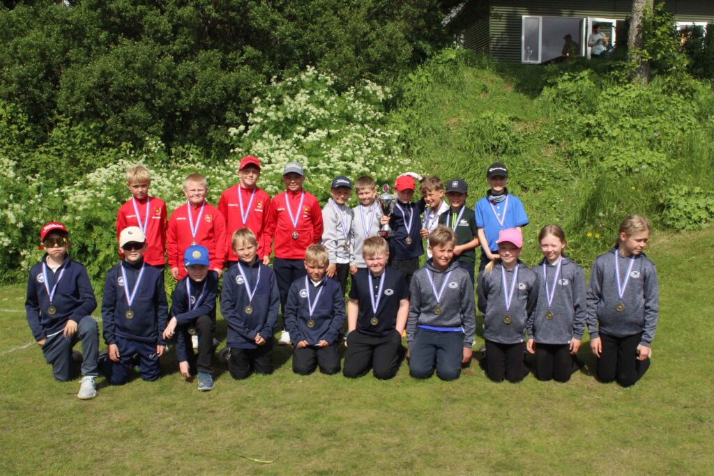 IMG_2866 1 - Golfsamband Íslands Group of children in sports uniforms posing on a grassy field with medals around their necks and a trophy in the center, outdoors on a sunny day.