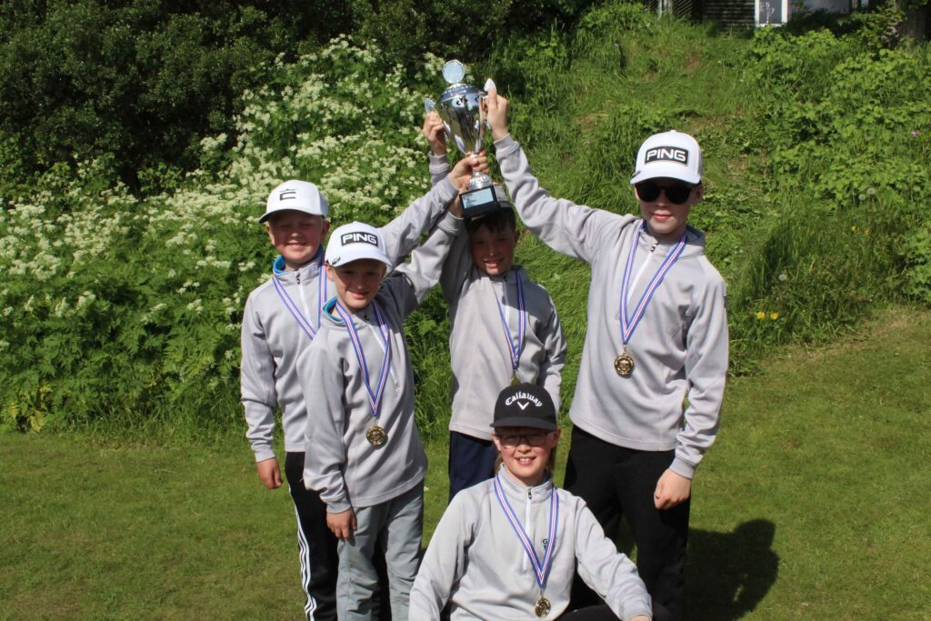 IMG_2870 1 - Golfsamband Íslands Group of young athletes in gray tracksuits with medals around their necks posing outdoors, lifting a large silver trophy in celebration.