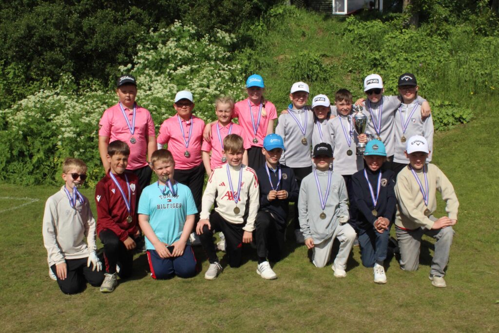 IMG_2872 1 - Golfsamband Íslands Group of kids outdoors posing with medals, some wearing pink shirts, and a trophy in a sunny grassy area.