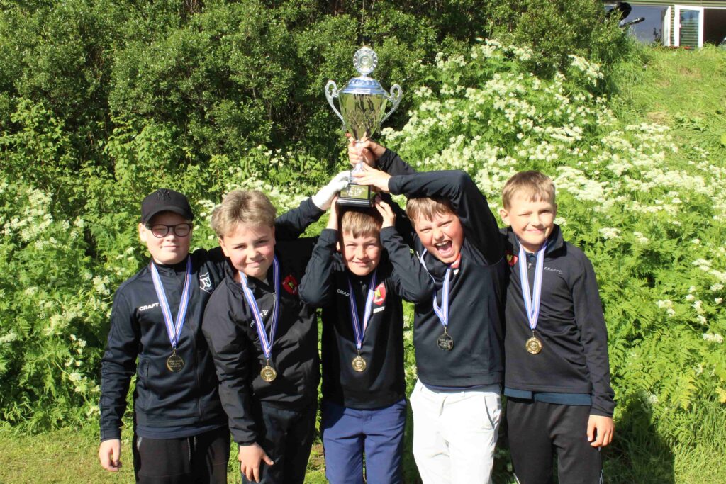 IMG_2878 1 - Golfsamband Íslands Group of young boys in dark jackets with medals around their necks, raising a large silver trophy outdoors in front of green bushes and white flowers, celebrating a win.