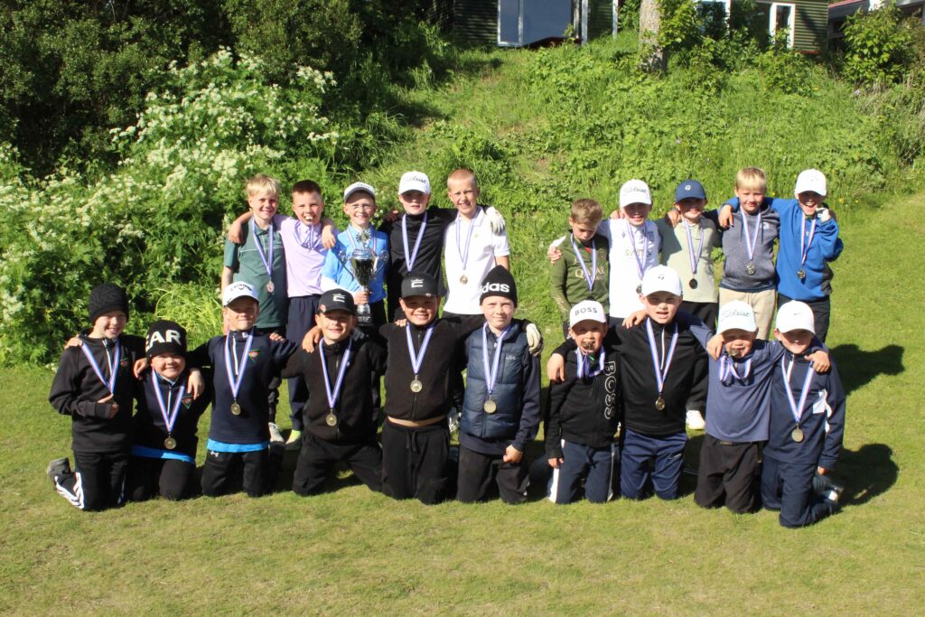 IMG_2884 1 1 - Golfsamband Íslands Group of young boys with medals posing outdoors after a sports event, a trophy held at center','Group of kids with medals standing in a line on a sunny grassy field with a trophy in the middle