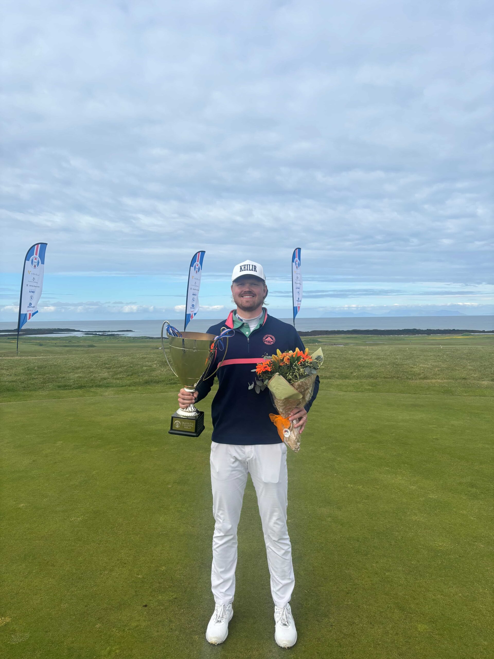Golfer standing on a course holding a large trophy in one hand and a bouquet of flowers in the other, with banners in the background and a coastal landscape behind him.