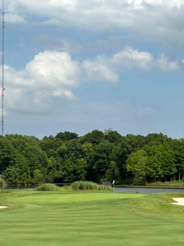 IMG_5662 - Golfsamband Íslands Green golf hole with flagstick on a well-manicured putting green, water and dense trees in the background, and a tall antenna tower to the left.
