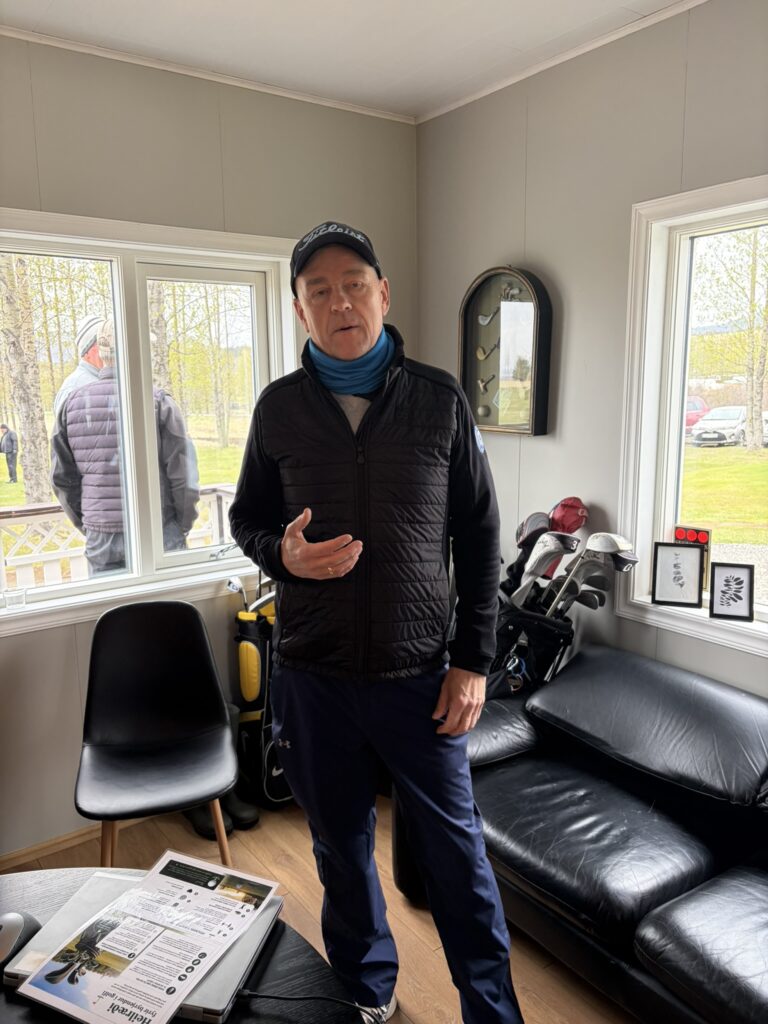 IMG_6043 - Golfsamband Íslands Man wearing a Titleist cap and blue neck gaiter, standing in a room with golf clubs and a black leather sofa.