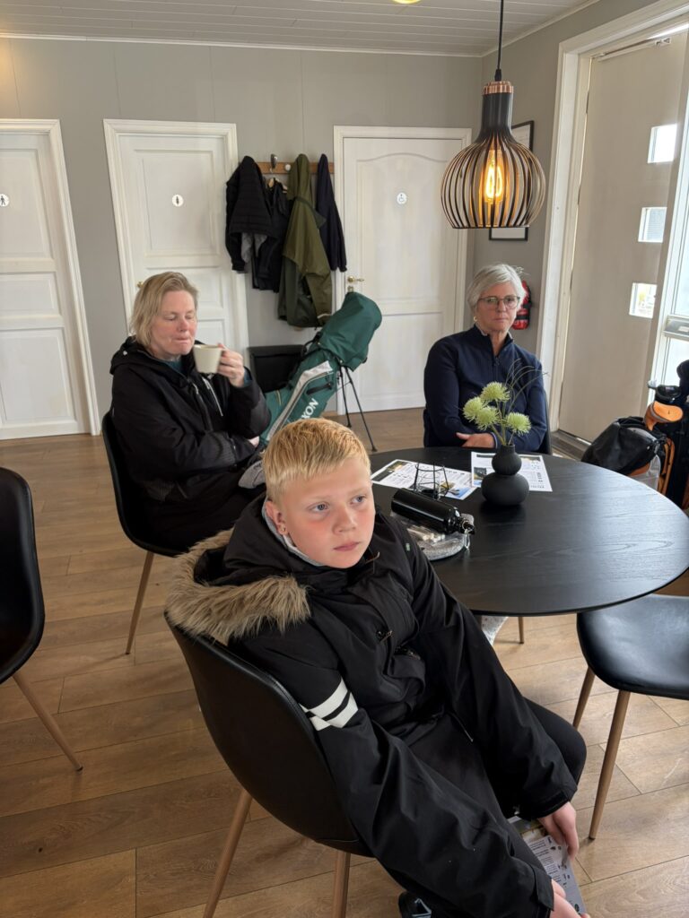 IMG_6044 - Golfsamband Íslands Child in a black parka with fur-trimmed hood sits at a round table in a cafe; two adults behind drink coffee and chat, coats hung on wall hooks.