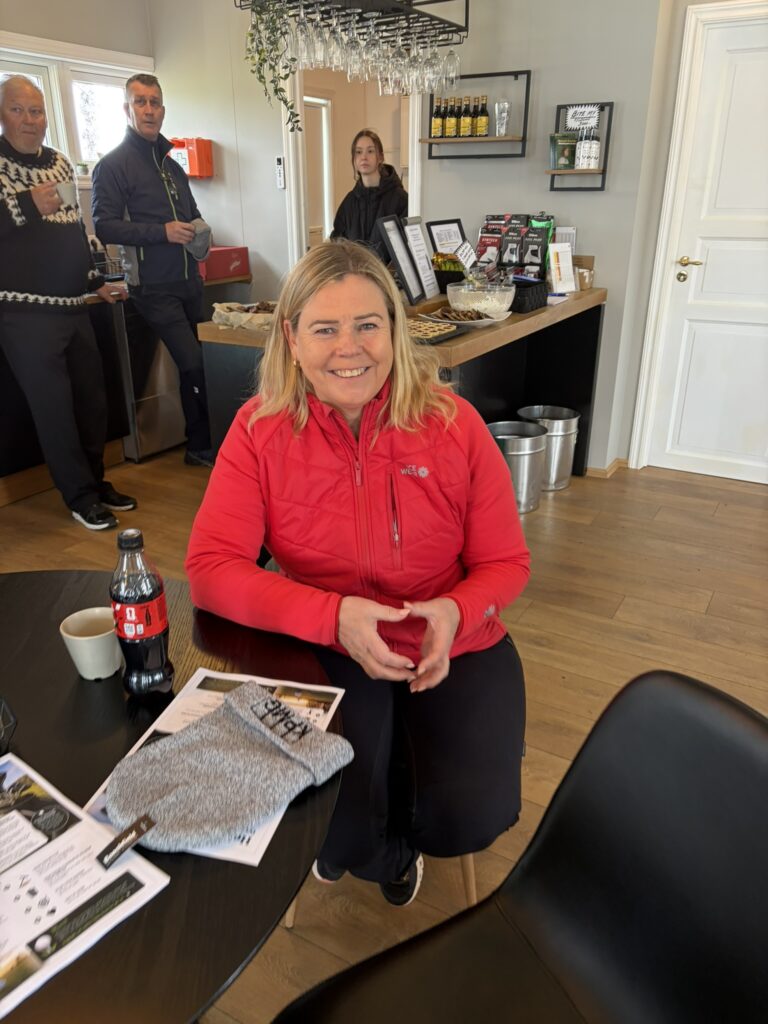 IMG_6045 - Golfsamband Íslands Smiling woman in a red jacket sits at a table in a cafe, with a Coke bottle and papers on the table nearby.