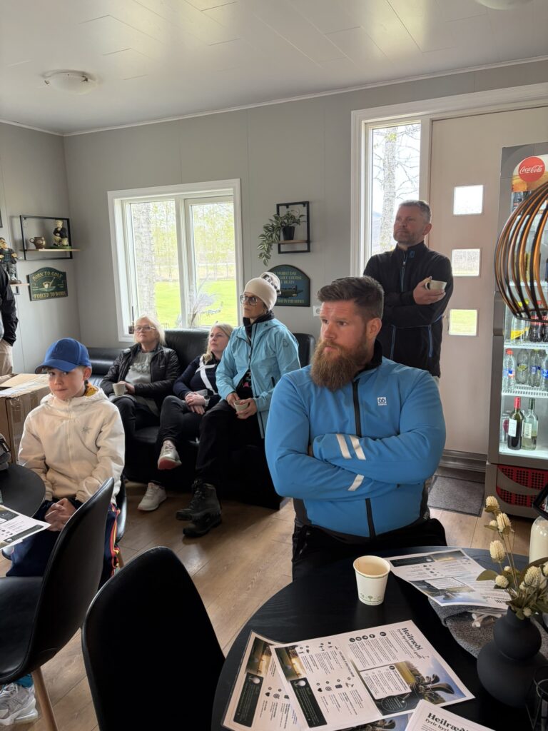 IMG_6046 - Golfsamband Íslands Group of people seated and standing in a cozy room, listening attentively during a small meeting, with coffee cups and papers on the table in the foreground.
