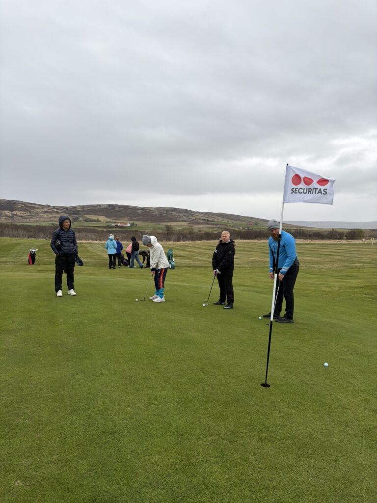 IMG_6049 - Golfsamband Íslands Group of people on a golf putting green outdoors, with a man holding a flag that says 'SECURITAS' while others prepare to putt.